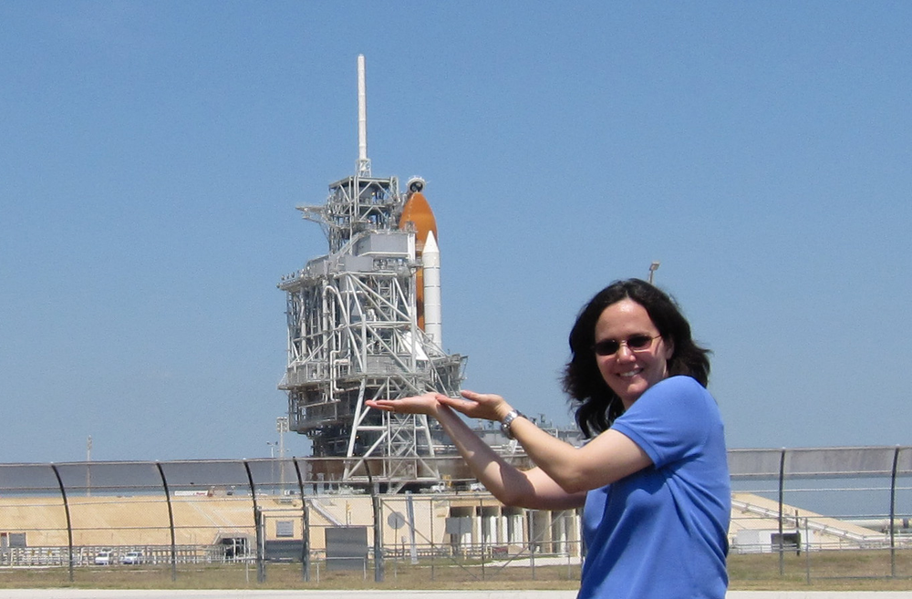 A woman in a blue shirt with a rocket on the launch pad behind her. 